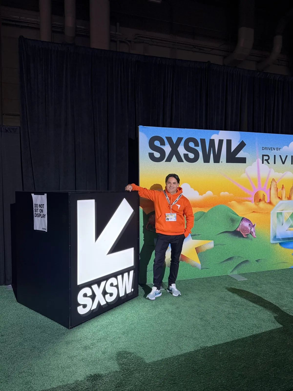 Ivaldo Trentin, de moletom laranja e crachá, ao lado do cubo luminoso com o logo do SXSW, em frente ao painel colorido do evento