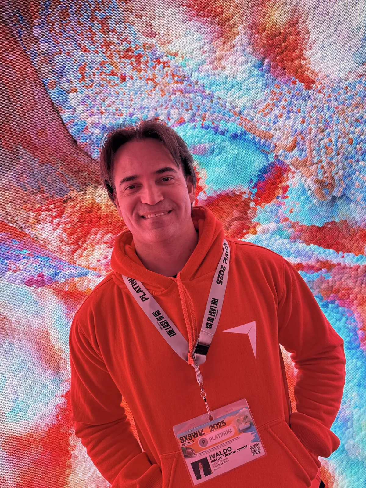 Ivaldo sorrindo diante de um painel de LED gigante com imagens coloridas, de moletom laranja e crachá do SXSW 2025
